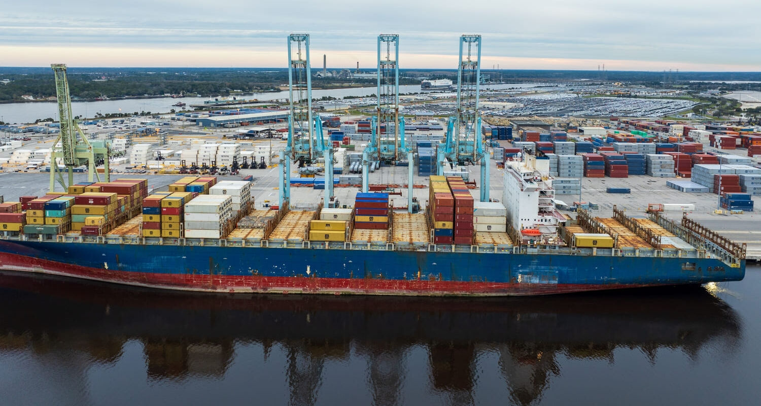 A container ship docked at Jacksonville port with cranes and stacked cargo containers, showing the city’s logistics and shipping hub.