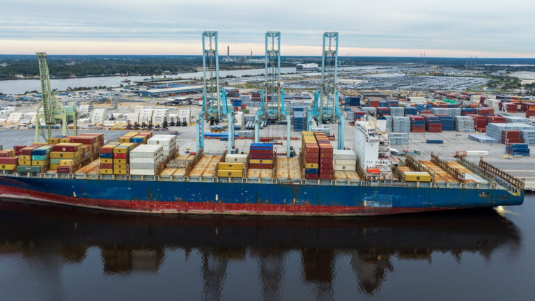 A container ship docked at Jacksonville port with cranes and stacked cargo containers, showing the city’s logistics and shipping hub.