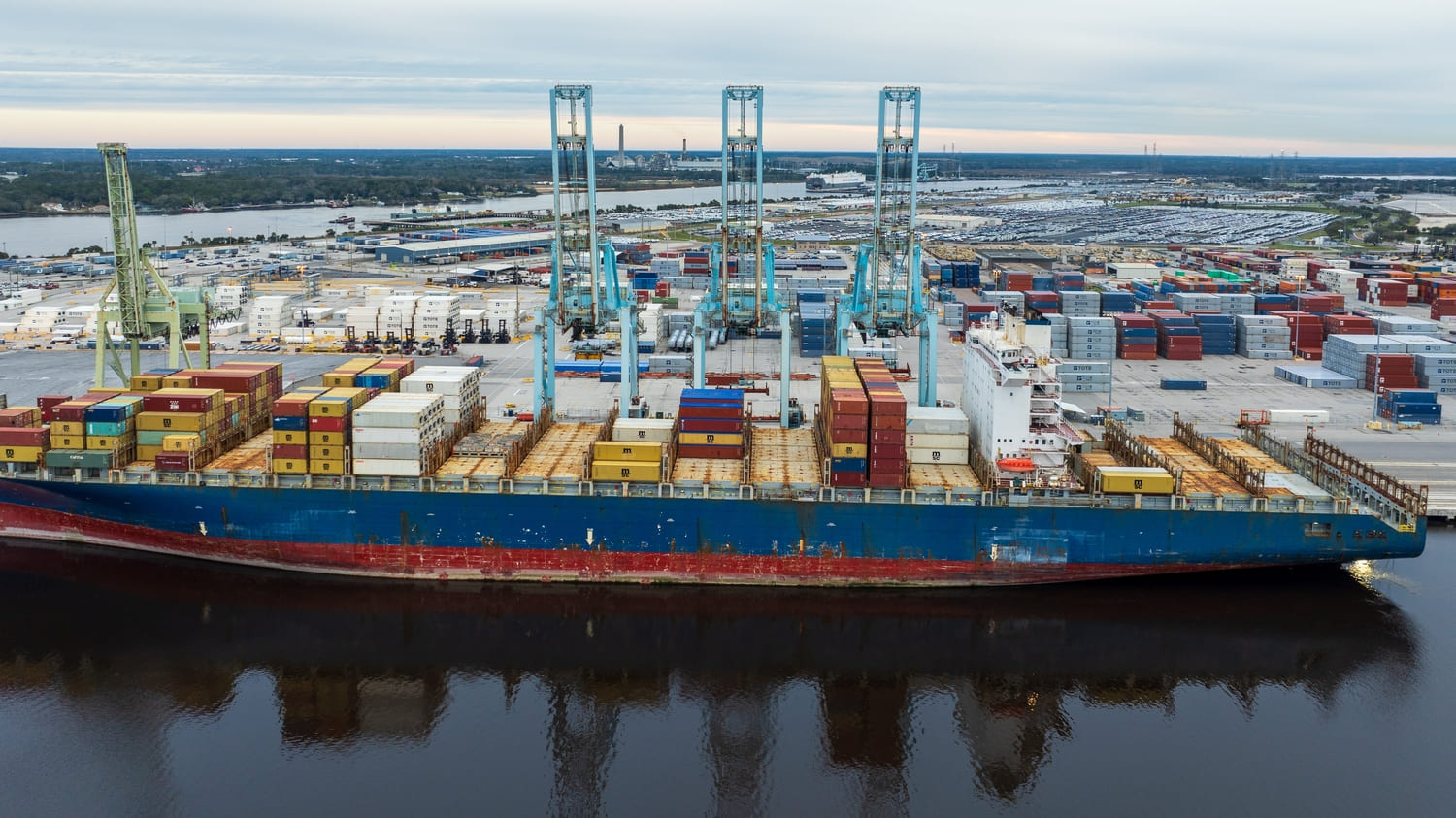 A container ship docked at Jacksonville port with cranes and stacked cargo containers, showing the city’s logistics and shipping hub.
